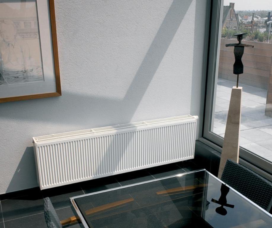 Просторная комната с панорамными окнами и низким стальным радиатором. Батарея белого цвета, стена светлая, рядом стоит обеденный стол и висит большая картина. Радиатор с нижним подключением и термоголовкой для удобного управления температурой