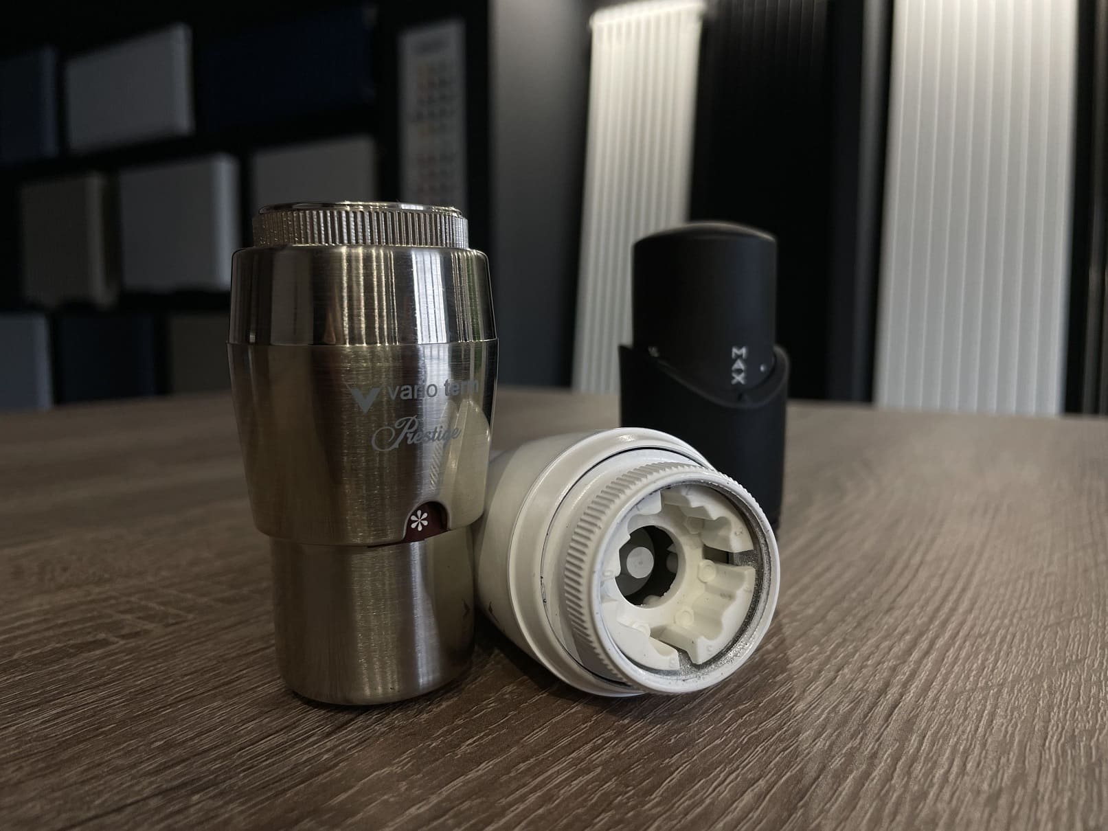 Термостатический клапан с регулируемой головкой в цвете хром, черный и белый. Они лежат на деревянном столе на фоне стенда с радиаторами. Поможет четко регулировать температуру радиатора или полотенцесушителя в помещении